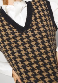 Hnědá a černá pletená vesta s houndstooth vzorem, s výstřihem do V a texturovaným povrchem, navrstvená přes bílou košilovou hranu.