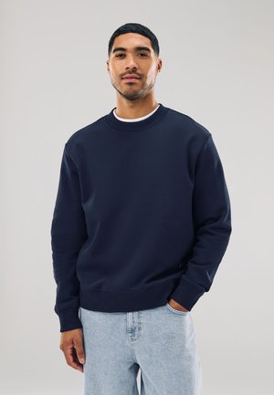 Jeune homme aux cheveux courts portant un sweat-shirt bleu marine et un jean bleu clair, debout avec une main dans la poche.
