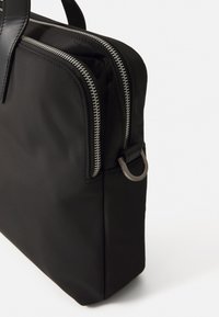 Bolsa de portátil negra de nailon con cremalleras dobles plateadas, forma rectangular plana, asas superiores y un lazo metálico de sujeción en un lado.