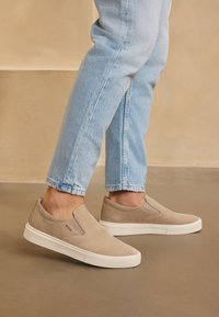 Slip-on tenisky v světlém béžovém semiši s bílou gumovou podrážkou. Disponují elastickými bočními vsadkami a hladkým, minimalistickým designem.