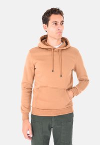 Giovane uomo con una felpa beige chiaro con tasca frontale e pantaloni verde scuro, che guarda di lato su uno sfondo neutro.