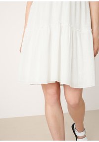 Robe midi blanche à étages portée avec des sandales blanches et noires à bouts ouverts, montrant les jambes et une partie des bras sur un fond neutre.