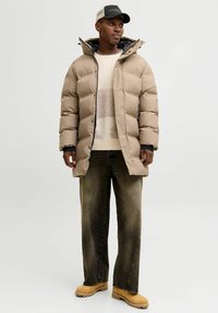 Mies pukeutunut beigenväriseen toppatakkiin, vaaleaan kuvioituun neuleeseen, haalistuneisiin mustiin farkkuihin, ruskeisiin saappaisiin ja mustaan baseball-lippikseen, seisoo valkoista taustaa vasten.