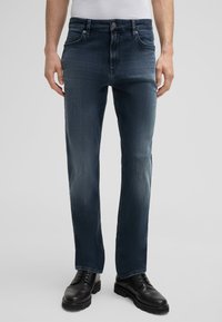 Dunkelblaue Jeans aus Denim mit geradem Bein, ausgestattet mit fünf Taschen und metallischen Elementen. Tragen mit schwarzen Lederschuhen.