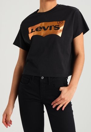 Osoba nosząca czarny krótki T-shirt Levi's z metalicznym miedzianym logo i czarne spodnie jeansowe z wysokim stanem, ręka na biodrze.