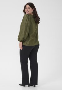 Nasmejana žena s dugom smešnom kosom nosi maslinasto zeleni blush s puf rukavima, crne pantalone i crne cipele s petom, stojeći naspram bele pozadine.