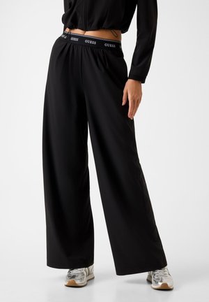 Donna con pantaloni neri a gamba larga con vita elastica con marchio Guess e sneakers argento, in piedi su sfondo semplice.