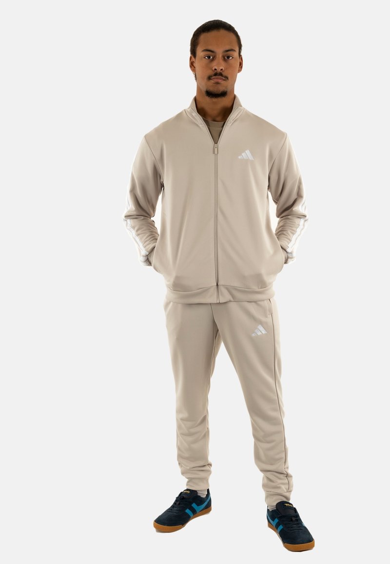 Completo giacca e pantaloni beige con accenti bianchi, frontale con zip e design a tre strisce sulle maniche. Sneakers nere e blu.