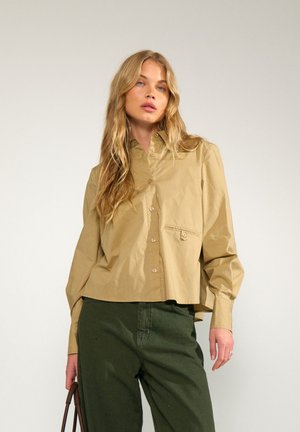 Femme aux longs cheveux blonds portant une chemise beige boutonnée et un pantalon vert foncé, tenant un sac à main marron à côté d’elle sur un fond uni.