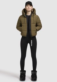 Giacca a vento verde olive con cappuccio, zip frontale e tasche laterali, abbinata a leggings neri e stivali foderati in pelliccia nera.