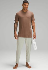 Bărbat cu tatuaje, îmbrăcat cu un tricou maro și pantaloni gri deschis, desculț, ținând o sticlă de apă albă Lululemon, pe un fundal gri simplu.