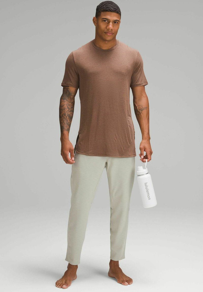 Bărbat cu tatuaje, îmbrăcat cu un tricou maro și pantaloni gri deschis, desculț, ținând o sticlă de apă albă Lululemon, pe un fundal gri simplu.