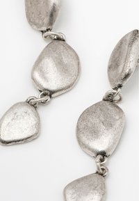 Orecchini in metallo tono argento con una forma organica e liscia, finitura opaca e collegamenti tra i singoli pezzi.