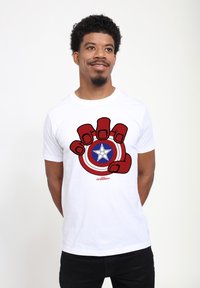 Hvid bomulds T-shirt med et grafisk motiv af en stor rød hånd, der griber om et cirkulært skjold med en blå stjerne og røde og hvide koncentriske cirkler.