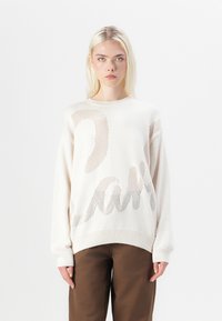 Krämfärgad stickad tröja med lös passform, med en texturerad, nätliknande design och ett djärvt grafiskt mönster i brunt över framsidan.