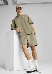Khaki tričko a šortky vyrobené z mäkkej tkaniny, s malým logom. Biele tenisky so zlatými akcentmi a ponožkami, postavené na bielom pozadí.