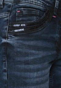 Vaqueros de mezclilla oscura con una textura suave, que cuentan con un bolsillo trasero adornado con lentejuelas negras y un pequeño detalle de costura rosa.