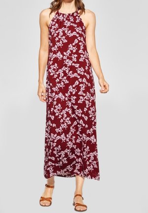 Robe maxi bordeaux avec un motif floral blanc, design à encolure halter, en tissu léger et longueur cheville. Portée avec des sandales brunes à lanières.