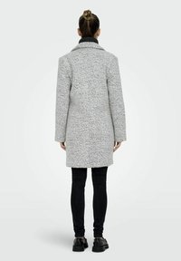 Grijze geweven jas met een gestructureerde afwerking, voorzien van een reverskraag en lange mouwen, gecombineerd met zwarte smalle broek en stevige zwarte schoenen.