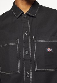 Chemise noire avec des coutures blanches, dotée d'un col, de boutons sur le devant, de deux poches poitrine et d'un petit patch logo Dickies multicolore.