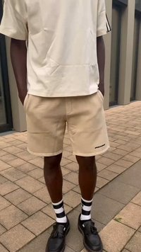 Bēša īsās bikses ar sānu kabatām, melna apzīmējuma, saskaņotas ar krēmkrāsas īsām piedurknēm, melnām kurpēm un melnbaltām svītrainām zeķēm.