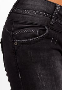 Jeans in denim nero con cuciture grigie a contrasto, dettagli invecchiati e decorazioni sulle tasche. Vista ravvicinata della parte sul retro e della vita.