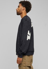 Zwarte sweatshirt met ronde hals, met een gestructureerde afwerking en grote witte afbeelding op de linkermouw. Randen zijn lichtjes rafelig.