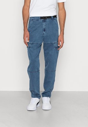 Mężczyzna ubrany w niebieskie dżinsowe spodnie cargo z bocznymi kieszeniami, czarny pasek, białe trampki i biała koszulka, stojący na szarej podłodze przy białej ścianie.