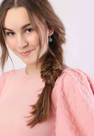 Modèle portant un t-shirt à côtes rose clair avec des manches bouffantes, des cheveux longs tressés et des boucles d'oreilles turquoises présentant des motifs circulaires.