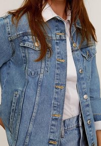 Světle modrá džínová bunda s klasickým designem, která má zapínání na knoflíky, dvě náprsní kapsy a zlaté kovové detaily.