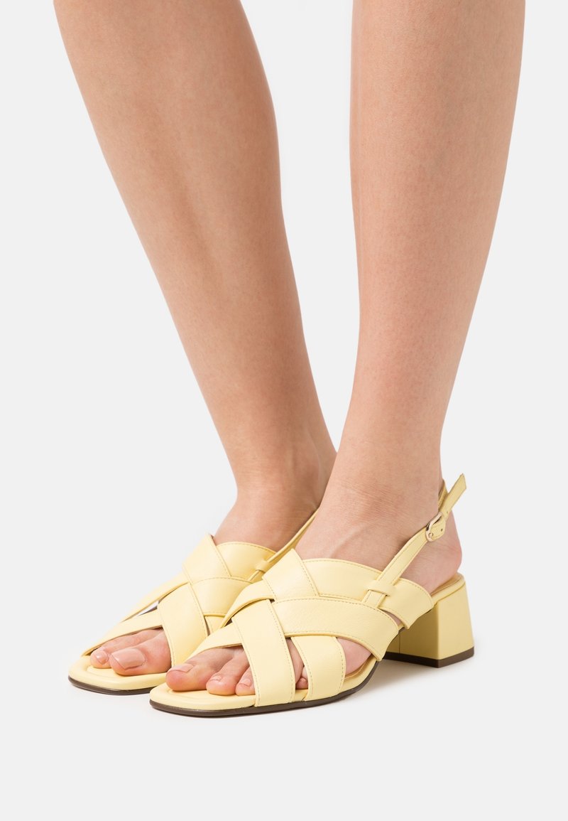 Pieds chaussés de sandales en cuir jaune pâle à talons carrés, avec des brides croisées et une boucle réglable à la cheville, sur un fond uni clair.