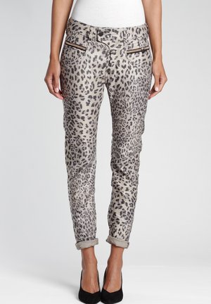 Femme portant un pantalon imprimé léopard retroussé avec des poches zippées à l'avant et des talons noirs, debout devant un fond uni.
