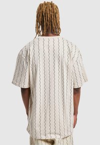 Krémové oversized tričko s vlnitým čiernym vzorom, krátkymi rukávmi, okrúhlym výstrihom, bavlnený materiál a voľný strih.
