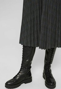 Botas de cuero negras hasta la rodilla con punta redondeada, suela plana y cordones verticales. Llevadas con una falda de cuadros gris plisada con acentos azules.