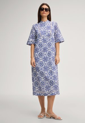 Vrouw draagt een midi-jurk met blauw-wit bloemenpatroon, ellebooglange mouwen, witte sandalen en zwarte oversized zonnebril.