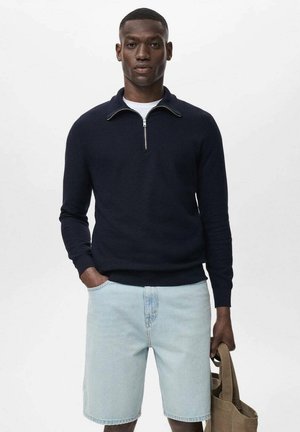 Marineblauer Strickpullover mit Reißverschlusskragen, kombiniert mit hellblauen Denim-Shorts. Das Model hält eine beige Tote-Tasche. Weiche Textur, lockere Passform.