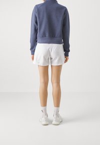 Sudadera polar azul con cuello alto, puños y dobladillo acanalados, combinada con pantalones cortos blancos. El modelo lleva calcetines blancos y zapatos de color claro.