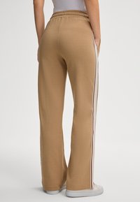 Helle beige Jogginghose mit lockerer Passform, ausgestattet mit seitlichen Streifen in Weiß und Kastanienrot. Elastischer Bund und seitliche Taschen für Funktionalität.