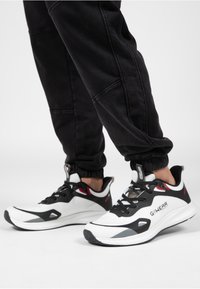 Witte en zwarte sportschoenen met rode accenten, voorzien van een gestructureerde bovenkant, vetersluiting en een flexibele, gevoerde zool.