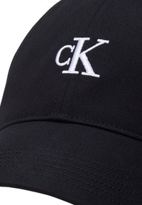Čierna baseballová čiapka s bielym vyšívaným logom "cK" na prednej strane, s oblúkovým šiltom a textúrovanou tkaninou.