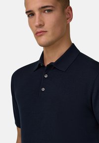 Jeune homme à la peau claire et aux yeux verts portant un polo bleu marine foncé à manches courtes avec deux boutons fermés.