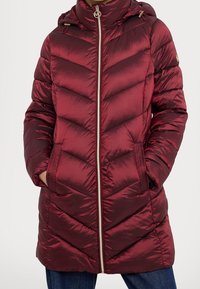 Viininpunainen puffatakki, jossa on viisarimuotoilu, vetoketjukiinnitys ja huppu. Varustettu kultasävyisillä yksityiskohdilla ja sivutaskuilla. Sileä, paljastettuna oleva rakenne.