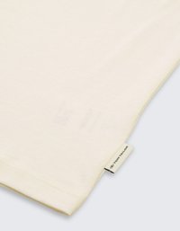 Cremfarbener Baumwoll-T-Shirt mit glatter Textur und kurzen Ärmeln, versehen mit einem Markenetikett am unteren Saum. Einfaches Design.