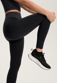 Crne legging hlače s visokim strukom, glatke teksture i detaljima na šavovima, uparene s crnim sportskim cipelama koje imaju teksturirani pleteni gornji dio i bijeli potplat.