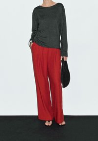 Grå langærmet top med løs pasform kombineret med røde bukser med vide ben. Sort taske holdt i den ene hånd, minimale accessories.