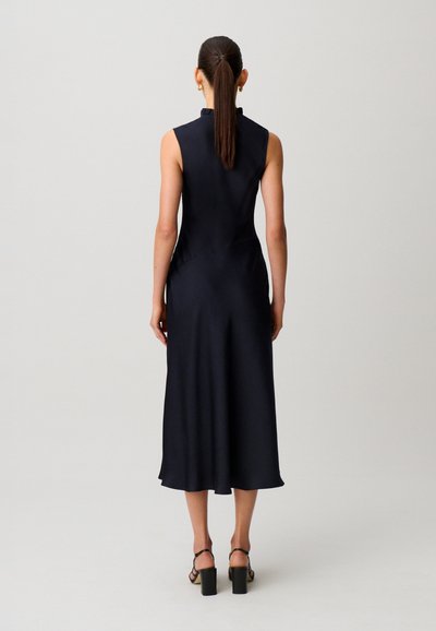 Mujer de pie vista de espaldas, con un vestido midi negro sin mangas, sandalias negras de tacón, cabello recogido en una coleta y pendientes dorados.