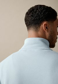 Lichtblauwe fleece pullover met een hoge kraag, een gladde textuur en minimale stikseldetails, gezien vanuit de achterkant.