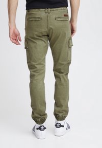Pantalons cargo vert olive avec des poignets élastiques, plusieurs poches et une texture lisse. Assortis à des baskets blanches avec des accents noirs.