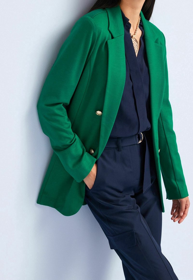 Personne portant un blazer vert sur un chemisier bleu marine et un pantalon taille haute assorti, les mains dans les poches, debout sur un fond clair.