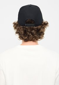Gorra de béisbol negra con un acabado texturizado, cierre ajustable tipo snapback y cabello visible por debajo; combinada con una camiseta blanca.